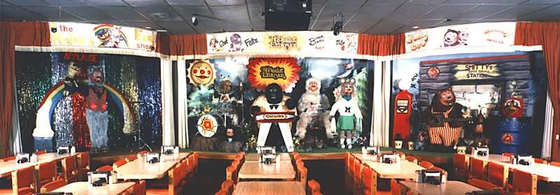 the rock-afire explosion animatronic band at showbiz pizza. the band is on three stages set before rows of booths, and all members are anthropomorphic animals. a wolf is stage right, on the left, holding a yellow puppet aloft in front of glittery streamers and an applause sign. center stage is made up to look like the outdoors. left to right is a dog on drums, a gorilla on keys, a polar bear on guitar sitting aside him, and a mouse with cheerleaders. on stage left, on the right, is a bear standing in front of a fake gas station with an oil barrel of goofy gas beside him, in which a red bird's head is visible, popping out.