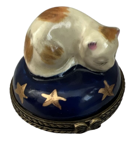 a figurine of a white and orange cat curled atop a jewelry box's top. the cat appears to be sleeping and the jewelry box is navy blue with gold stars like the night sky.